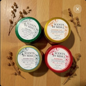 May include: Four round, handmade soy and coconut wax candles in tins. The tins are green, gold, gold, and red. Each tin has a label with the text "SCENTS BY MISS J." and a different scent name: Spearmint, Lemorange Cock, Lavender Velvet, and Raspberry.