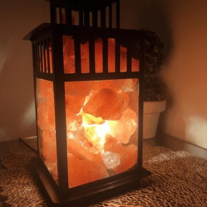 K&ouml;nnte beinhalten: Eine schwarze Metalllaternenlampe, gef&uuml;llt mit leuchtenden rosa Himalaya-Salzsteinen. Die Lampe hat eine rechteckige Form mit quadratischer Basis und dekorativer Oberseite. Das warme Licht erzeugt ein gem&uuml;tliches Ambiente.
