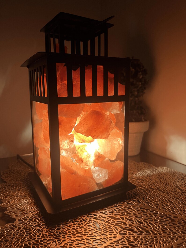 K&ouml;nnte beinhalten: Eine schwarze Metalllaternenlampe, gef&uuml;llt mit leuchtenden rosa Himalaya-Salzsteinen. Die Lampe hat eine rechteckige Form mit quadratischer Basis und dekorativer Oberseite. Das warme Licht erzeugt ein gem&uuml;tliches Ambiente.