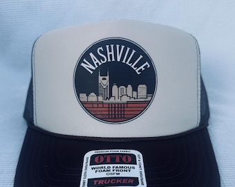 May include: A trucker hat with a black brim and mesh back, and a tan front panel. The front panel features a circular design with the word "Nashville" and a skyline graphic. The hat includes a tag that reads "OTTO Trucker".