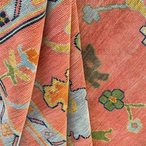 May include: Close-up of a folded coral-colored rug with a detailed floral pattern. The design incorporates light blue, yellow, and dark blue accents. The rug's texture is visible, showcasing its woven construction.