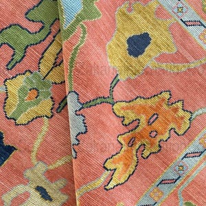 May include: Close-up of a section of a vintage rug with a floral pattern. The rug features a salmon-colored background with yellow, blue, green, and orange floral designs. The rug's texture is visible, suggesting a woven construction.