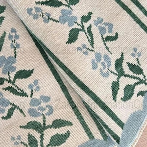 Op de afbeelding: Close-up van een beige tapijt met een bloemenpatroon. Het tapijt heeft lichtblauwe bloemen en groene bladeren op een beige achtergrond. Groene strepen en een lichtblauwe rand omlijsten het ontwerp. Het tapijt is opgevouwen en toont de textuur.