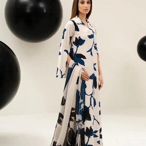 Puede incluir: Vestido largo color crema con estampado floral en azul marino y negro. El vestido presenta un cuello camisero, mangas acampanadas y una bufanda a juego. La modelo lleva sandalias negras. Dos grandes esferas negras en el fondo.