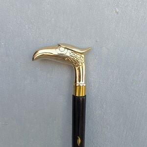 Könnte beinhalten: Dekorative Gehstock mit einem Griff in Form eines Adlerkopfes aus Messing. Der Griff ist goldfarben und der Schaft ist schwarz mit einem goldenen Akzent. Der Stock ist ein stilvolles Accessoire.