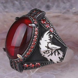 May include: A silver ring with a large, oval, deep red gemstone. The ring is embellished with smaller red stones and a detailed design on the band, including a white animal head.