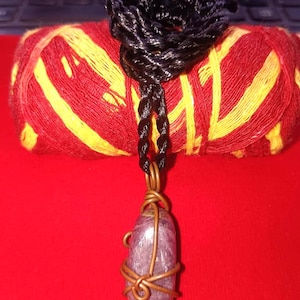 Puede incluir: Un collar con colgante hecho a mano con una piedra púrpura ovalada pulida, envuelta en alambre de cobre. El collar cuelga de un cordón negro. El fondo incluye una madeja de hilo rojo y amarillo.
