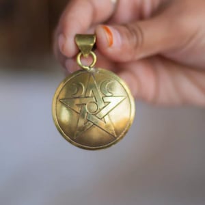 Peut inclure: Un pendentif doré avec un motif de pentagramme, comprenant une étoile centrale et des accents de croissant de lune. Le pendentif est de forme circulaire et possède une boucle pour une chaîne. Le design général est détaillé et complexe.