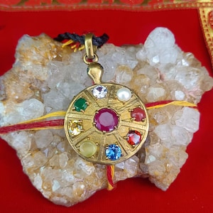 May include: A gold pendant with a circular design, featuring a large ruby in the centre and smaller gemstones of various colours around it. The pendant is displayed on a rough, light-coloured stone against a red background.