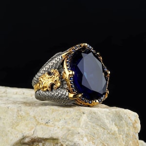 May include: A large, ornate ring featuring a faceted, deep blue gemstone. The ring has a gold-colored bezel and accents, with smaller blue stones surrounding the main stone. The band is silver-colored with a textured pattern and gold filigree.