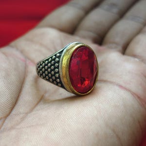 Pode incluir: Um anel com uma grande pedra vermelha facetada de formato oval, engastada num bisel dourado. A banda é feita de metal prateado oxidado e apresenta um padrão texturizado de pontos nas laterais, criando um contraste com a pedra lisa.
