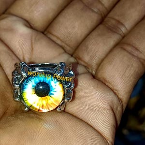 Könnte beinhalten: Ein silberfarbener Ring mit einem großen, farbenfrohen Augendesign. Das Auge hat eine blaue Iris mit gelben und orangen Akzenten, und der Text "worship power" ist sichtbar. Der Ring wird in einer Hand gehalten.