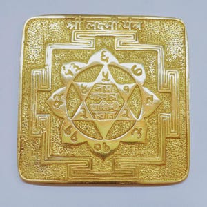 Op de afbeelding: Een goudkleurige vierkante plaat met ingewikkelde reliëfontwerpen. De centrale focus is een zespuntige ster in een lotusachtige vorm, omringd door geometrische patronen en cijfers. De bovenkant van de plaat heeft tekst in een vreemd schrift.