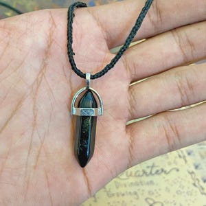 May include: Black crystal pendant necklace with a silver-colored metal bail and a black cord. The crystal is a dark, elongated hexagonal shape. The necklace is held in a hand.