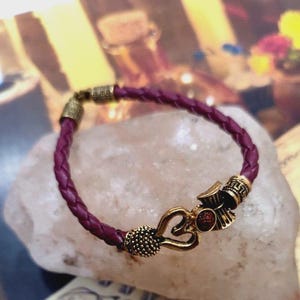 May include: A braided burgundy leather bracelet with gold-colored metal accents. The bracelet features a heart-shaped clasp and decorative end pieces. The bracelet is displayed on a light-colored, textured surface.