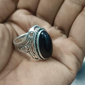 May include: A silver ring featuring a large, oval black gemstone. The ring has an intricate pattern on the band and around the stone. The gemstone is polished. The ring is held in a hand.