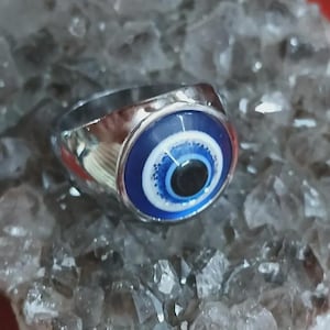 May include: A silver-toned ring featuring a blue and white eye design with a black pupil. The ring has a polished finish and is set against a textured, crystal-like background. The eye design is a prominent focal point.
