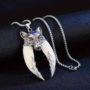 Może przedstawiać: Srebrny naszyjnik z wisiorkiem w kształcie głowy wilka z czerwonymi oczami i niebieskim kamieniem. Wisiorek posiada duże, białe kły ze srebrnymi, płomienistymi akcentami. Łańcuszek naszyjnika jest srebrny.