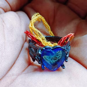 May include: A dark ring with a blue heart-shaped gemstone. The ring has a textured, melted-like design and is adorned with yellow and red threads. The ring is held in a hand.