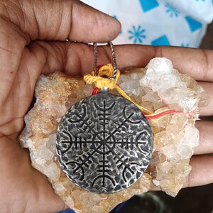 Könnte beinhalten: Ein silberfarbener Anhänger mit einem detaillierten, symmetrischen Design, der an einer filigranen Kette hängt. Der runde Anhänger wird vor einem rauen, hellfarbigen Stein präsentiert. Die Kette ist mit gelben und roten Fäden befestigt.