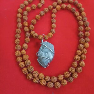 May include: A mala necklace with brown beads and a silver-wrapped stone pendant. The beads are strung on a pink cord. A yellow and orange tassel hangs from the top of the necklace. The necklace is displayed on a red surface.