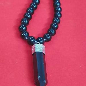 May include: A black beaded necklace featuring a black, hexagonal crystal pendant with a silver-coloured metal cap. The necklace is set against a red background.