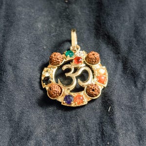 Puede incluir: Colgante dorado con el símbolo Om, adornado con piedras preciosas de colores y cuentas marrones. El colgante tiene un lazo para una cadena y está sobre un fondo oscuro.