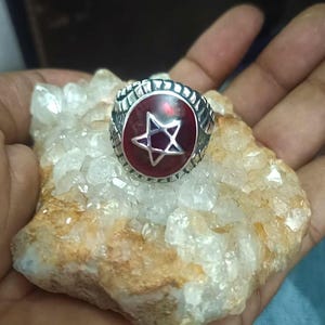 Może przedstawiać: Srebrny pierścionek z czerwonym środkiem z białym wzorem gwiazdy. Pierścionek osadzony jest na chropowatym, jasnym kamieniu. Opaska pierścionka ma teksturowany wzór.