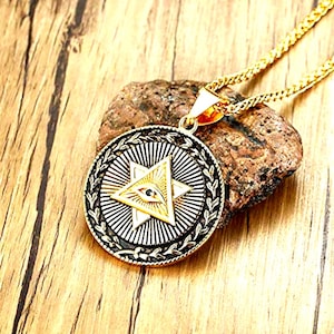 Może przedstawiać: Złoty naszyjnik z wisiorkiem z okrągłym wzorem z symbolem gwiazdy i oka. Wisiorek ma czarno-białe detale i złoty łańcuszek. Naszyjnik jest prezentowany na drewnianej powierzchni.