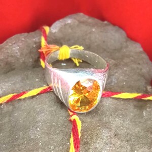 Może przedstawiać: Srebrny pierścionek z dużym, fasetowanym, żółto-pomarańczowym kamieniem szlachetnym. Pierścionek leży na szarej skale, z czerwonymi i żółtymi nićmi wokół niego. Tło jest jednolicie czerwone.