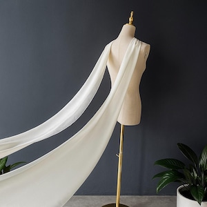 May include: A cream-colored fabric draped over a mannequin, creating a flowing effect. The fabric is lightweight and appears to be silk or a similar material. The mannequin is on a gold stand, set against a dark gray background.