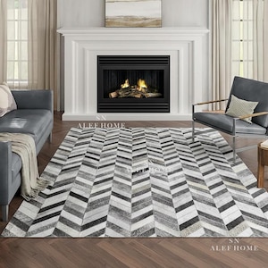 May include: A large, rectangular area rug with a chevron pattern in shades of gray, black, and white. The rug is made of a textured material and is placed on a hardwood floor. The rug is in a living room setting.