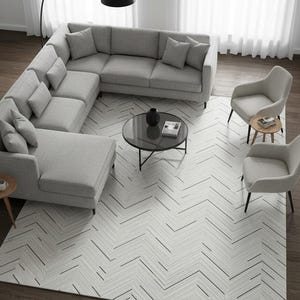 Op de afbeelding: Een bovenaanzicht van een moderne woonkamer met een grijze hoekbank, twee fauteuils en een ronde salontafel. Een patroonkleed met zwarte lijnen bedekt de vloer. Een zwarte booglamp en bijzettafels maken de inrichting compleet.