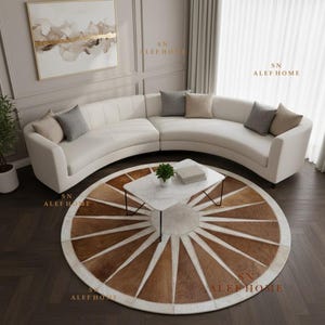 Può includere: Un grande divano curvo color crema con diversi cuscini grigi e beige, posizionato dietro un tavolino rettangolare bianco con piano effetto marmo, il tutto poggiato su un tappeto circolare con un motivo a raggiera marrone e bianco.