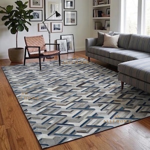 Op de afbeelding: Een rechthoekig vloerkleed met een geometrisch patroon van in elkaar grijpende lijnen in tinten grijs, wit en marineblauw. Het kleed heeft een laagpolige textuur en ligt op een houten vloer in een kamer met een bank, stoel en plant.