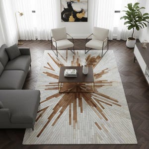 May include: A contemporary living room with a striking area rug. The rug has a starburst pattern in brown and white. The room also features a grey sofa, two cream armchairs, and a dark brown coffee table with a gold frame.