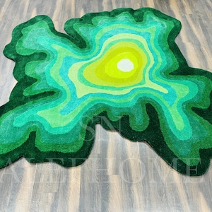 May include: A green, abstract rug with a topographic design. The rug features concentric layers of varying shades of green, from dark forest green to a bright yellow-green center. The rug's shape is irregular, resembling a map.