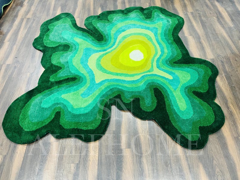 May include: A green, abstract rug with a topographic design. The rug features concentric layers of varying shades of green, from dark forest green to a bright yellow-green center. The rug's shape is irregular, resembling a map.
