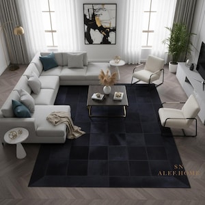 Könnte beinhalten: Ein Innenraum eines modernen Wohnzimmers mit einem großen grauen Ecksofa, zwei weißen Sesseln und einem dunkelblauen quadratisch gemusterten Teppich. Ein abstraktes Gemälde in Schwarz und Gold hängt an der Wand. Der Raum ist hell.