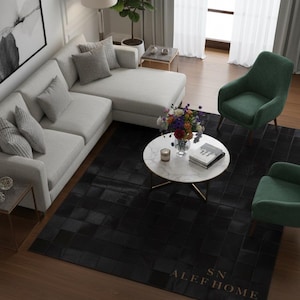 Può includere: Un moderno soggiorno con un divano componibile grigio chiaro, due poltrone verdi e un tavolino rotondo in marmo. Un tappeto nero con un motivo geometrico copre il pavimento. Il tappeto ha il testo "SN ALEF HOME" in oro.