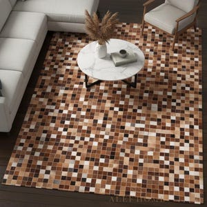 Könnte beinhalten: Ein rechteckiger Teppich mit einem Mosaikfliesenmuster in Braun-, Beige- und Weißtönen. Der Teppich liegt auf einem dunklen Holzboden, mit einem weißen Ecksofa, einem runden Marmor-Couchtisch und einem beigen Sessel im Hintergrund. Der Teppich hat den Text "SN ALEF HOME".