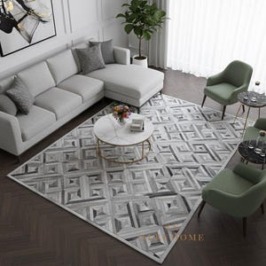 May include: A modern living room setup featuring a light gray sectional sofa, a geometric patterned rug, and two green armchairs. A round marble coffee table with a gold base sits in the center. The room has hardwood floors and a large window.