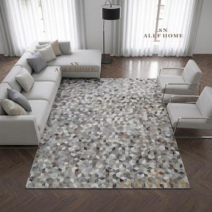 Op de afbeelding: Een groot rechthoekig vloerkleed met een geometrisch kubuspatroon in grijze, witte en bruine tinten. Het kleed ligt op een donkere houten vloer in een kamer met een lichtgrijze hoekbank, twee fauteuils en een zwarte vloerlamp.