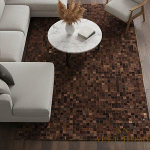 May include: A brown and tan square-patterned rug dominates the room. A white sectional sofa and a matching armchair surround a white marble coffee table. The rug is made of small, square tiles in varying shades of brown. The floor is wooden.
