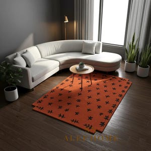 May include: A modern living room setup featuring a curved, light gray sofa, a small round coffee table, and an orange rug with a black floral pattern. Potted plants and a floor lamp add to the decor. The room has a neutral color palette.