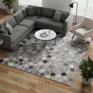 Op de afbeelding: Een moderne woonkamer met een grijze hoekbank, een witte fauteuil en een ronde salontafel. Een geometrisch patroon tapijt met zeshoekige vormen in grijstinten en zwart bedekt de vloer. Decoratieve planten voegen een vleugje natuur toe.