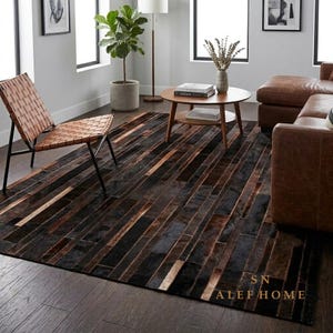 Può includere: Un tappeto rettangolare con un design patchwork composto da lunghe strisce strette in varie tonalità di nero, marrone scuro e marrone chiaro. Il tappeto è posizionato in una stanza con sedia in pelle, tavolino in legno e divano. Testo: SN ALEF HOME.