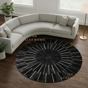 May include: A round area rug featuring a sunburst pattern with alternating black and metallic silver panels. The rug is positioned on a dark wood floor in front of a curved light grey sofa. The text SN ALEF HOME is visible on the rug surface.