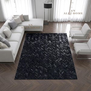 May include: A rectangular area rug featuring a dark black and charcoal grey chevron pattern composed of individual leather tiles. The rug is positioned on a dark brown herringbone wood floor in a room with a white sectional sofa and two chairs.