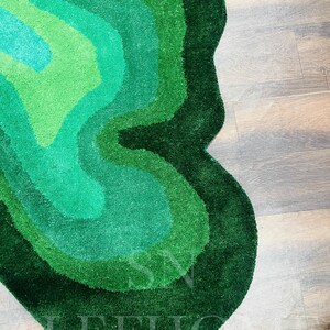 May include: A green, abstract-shaped rug with varying shades of green, from light to dark. The rug has a soft, textured surface and is placed on a wooden floor. The design features layered, organic shapes.